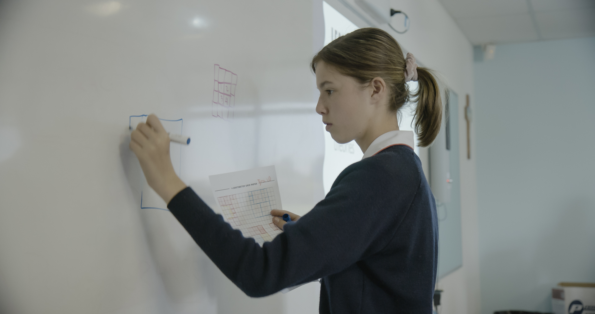 One girl draws a shape on the whiteboard, her face intense with focus. 